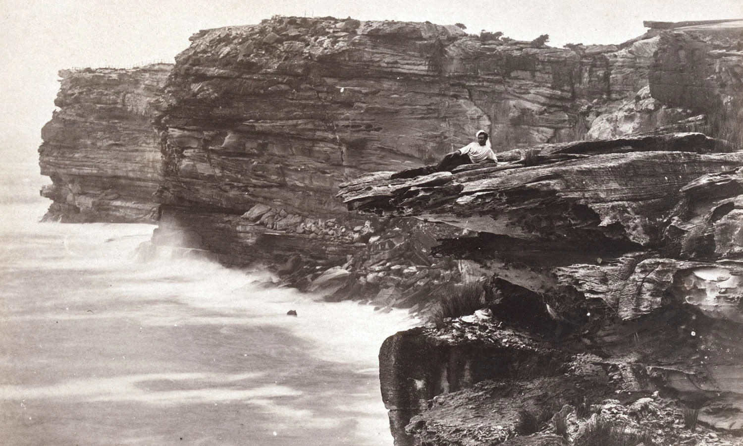 Early photograph of a man reclining on a cliff edge overlooking the ocean.