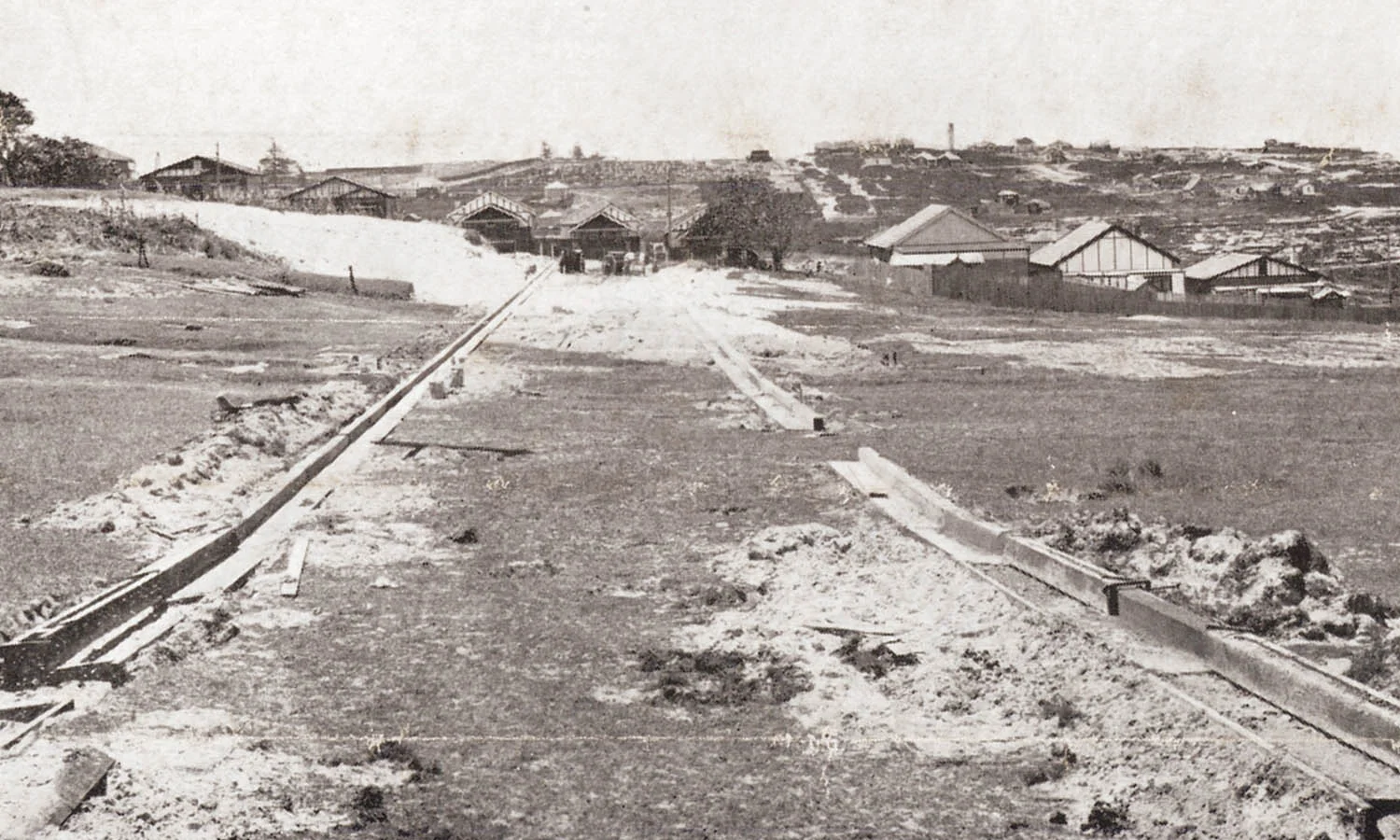 An old black and white photo depicting a road being made. There are houses in the distance.